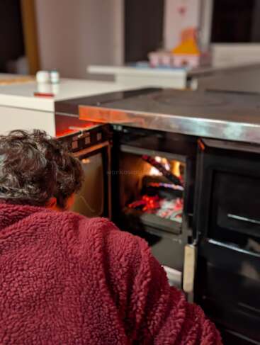 Eine Person in einer roten Fleecejacke kniet vor einem offenen Holzofen und kümmert sich um ein Feuer in einer gemütlichen Küche.