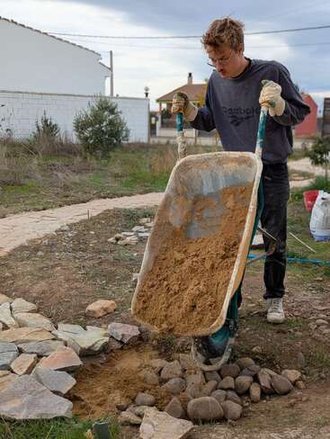Ein junger Mann mit Handschuhen und einem Pullover schüttet Sand aus einer Schubkarre auf Steine. Er arbeitet im Freien und baut oder repariert möglicherweise einen Steinweg in einem Garten.