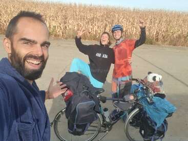Drei glückliche Freunde posieren zusammen an einem vollgepackten Tourenrad, lächeln und feiern draußen in der Nähe eines Maisfeldes. Ein ausgestopfter Panda sitzt auf dem Fahrrad.
