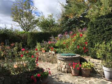 Una encantadora escena de jardín con senderos de piedra, un pozo circular de piedra, coloridas flores en flor, follaje verde, plantas en macetas y árboles bajo un cielo parcialmente nublado.