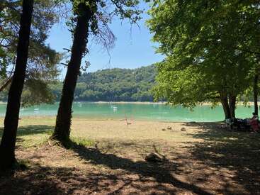 Una tranquila escena junto al lago con aguas turquesas, a la sombra de altos árboles. Hay colinas al fondo, un columpio cerca de la orilla y gente relajándose.