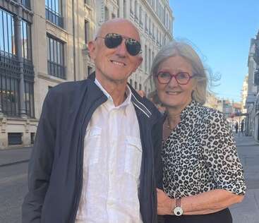 Una pareja de ancianos está de pie, muy juntos, en una soleada calle de la ciudad. El hombre lleva gafas de sol y chaqueta; la mujer lleva gafas rojas y una blusa estampada.