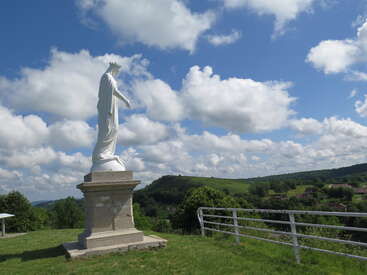 Una estatua blanca de una figura coronada se erige sobre un pedestal con vistas a un frondoso y verde valle, bajo un cielo lleno de nubes esponjosas y una brillante luz solar.