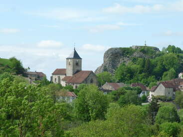 A charming village features a stone church with a clock tower, surrounded by lush greenery, houses, and a rocky hill topped with a statue. Peaceful scene.