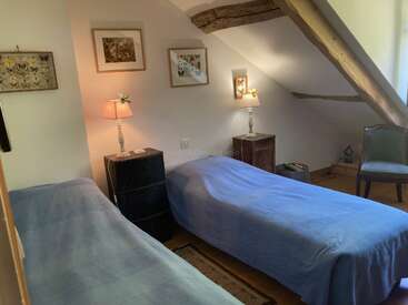This cozy attic bedroom features two single beds with blue covers, wooden beams on the ceiling, framed art on the walls, lamps, and a green chair.