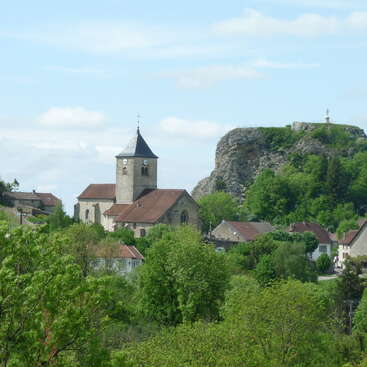 A charming village features a stone church with a clock tower, surrounded by lush greenery, houses, and a rocky hill topped with a statue. Peaceful scene.
