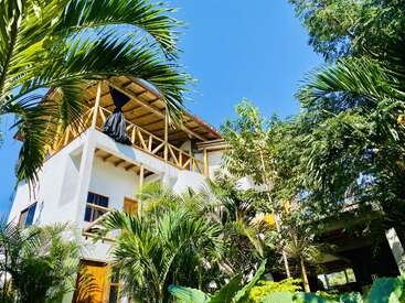 A modern, white, multi-story tropical house surrounded by lush greenery and palm trees, featuring wooden balcony railings and a thatched roof under a bright blue sky.