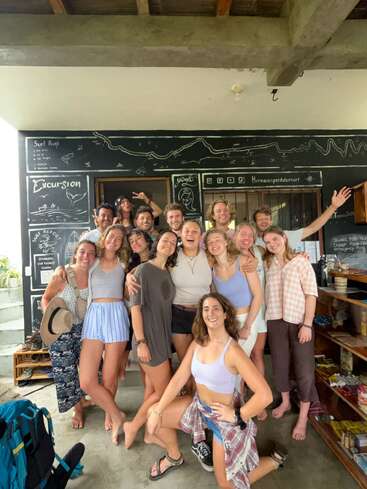 A group of fifteen smiling young adults poses together indoors, barefoot and casual, in front of a chalkboard wall covered with surf-related drawings and notes.