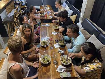 Eight people sit around a wooden table, enjoying bowls of food, chatting, and sharing a meal in a cozy, casual restaurant with a warm, inviting atmosphere.
