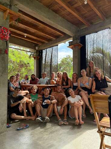 A group of friends gathers indoors, sitting and smiling together. Sunlight streams through windows, creating a warm, happy, relaxed atmosphere. Plants and wooden accents decorate the space.