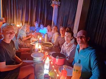 A group of people sit around a table in a warmly lit room, sharing food, drinks, and laughter, creating a cozy, joyful atmosphere filled with friendship.