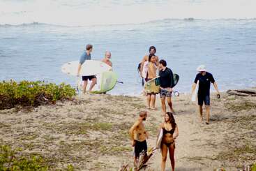 A group of people, some holding surfboards, walk along a sandy beach near the ocean. They appear happy, chatting, and enjoying a sunny day together.