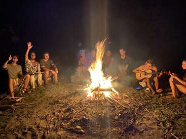 A group of friends sit around a glowing campfire at night, some waving, others playing guitar, enjoying music and conversation under the open, dark sky.