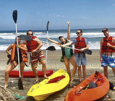 The image shows five people standing on a beach, each holding a kayak paddle and wearing life jackets, with kayaks scattered around them.