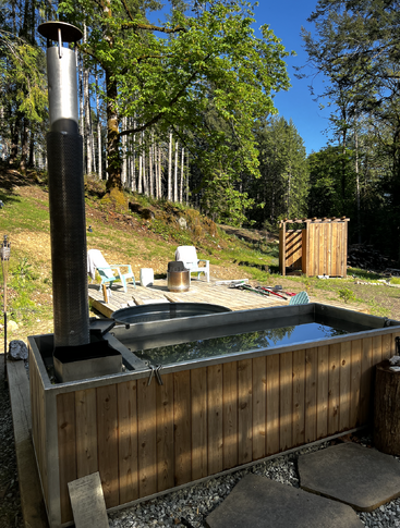 A rustic outdoor hot tub made of wood and metal sits in a forested area, with lounge chairs, a fire pit, and a wooden privacy screen nearby.