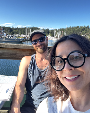 A smiling couple enjoys a sunny day by a marina. Boats and yachts are docked on the water, with a forested background and clear blue sky.