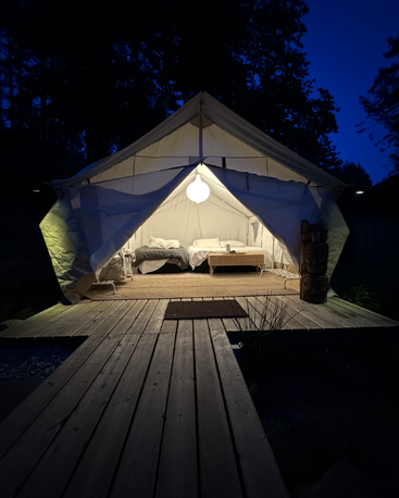 A cozy glamping tent glows warmly at night, nestled among trees. Inside, two beds and a wicker table invite restful comfort and relaxation in nature.