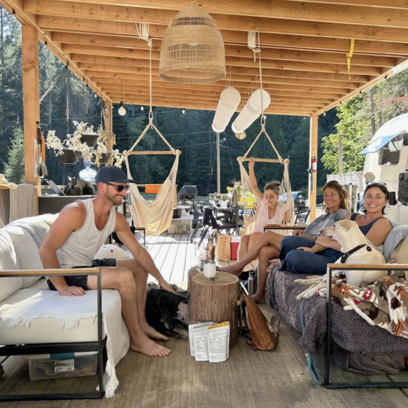 Four people relax under a wooden outdoor patio, chatting and smiling. Two dogs join them. The setting is cozy, surrounded by trees, hammocks, and natural light.