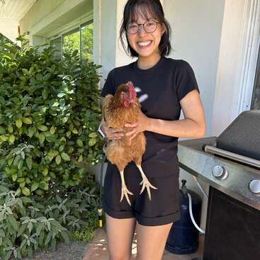 Una mujer joven con el pelo oscuro y gafas está de pie fuera, sosteniendo un gran pollo marrón, vestida con una camiseta negra y pantalones cortos, delante de una casa y una parrilla.