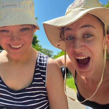 Two young women smile brightly outdoors on a sunny day, both wearing wide-brimmed hats. They seem happy and energetic, enjoying nature and each other's company.