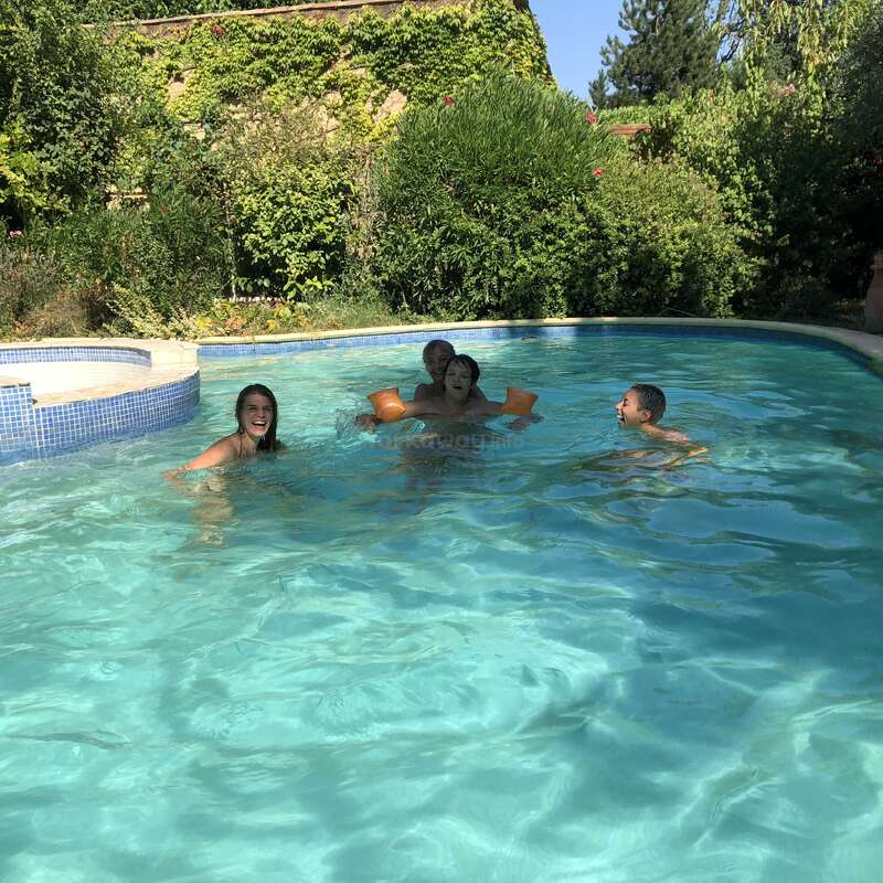 L'image représente une scène de piscine sereine avec quatre personnes nageant et jouant dans l'eau, entourées d'une végétation luxuriante et d'un ciel bleu clair.