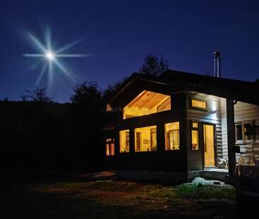 Una acogedora casa brilla cálidamente por la noche, rodeada de oscuridad. La luna brillante y estrellada brilla en el cielo azul despejado, iluminando la tranquila y apacible escena.