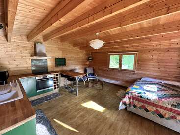 El interior de esta acogedora cabaña de madera cuenta con una pequeña cocina, un comedor y una cama con una colcha de colores. La luz natural inunda el espacio a través de una ventana.