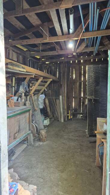 This image shows a rustic wooden shed with a dirt floor, exposed beams, shelves filled with tools and materials, and blue pipes attached to the ceiling.