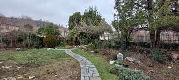 Ein Garten mit einem Steinweg schlängelt sich durch Gras und Bäume. Bewölkter Himmel, verschiedene Sträucher und Pflanzen, vereinzelte Töpfe und ein Drahtzaun im Hintergrund.