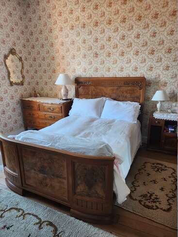 Dieses gemütliche Schlafzimmer im Vintage-Stil besticht durch ein Holzbett, eine Blumentapete, eine antike Kommode und einen Spiegel, Spitzenlampen sowie passende Teppiche, die eine warme, nostalgische Atmosphäre schaffen.