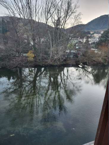 Am Flussufer stehen kahle Bäume, deren Äste sich wunderschön im ruhigen Wasser spiegeln. Im friedlichen Hintergrund sind Hügel und ein kleines Dorf zu sehen.