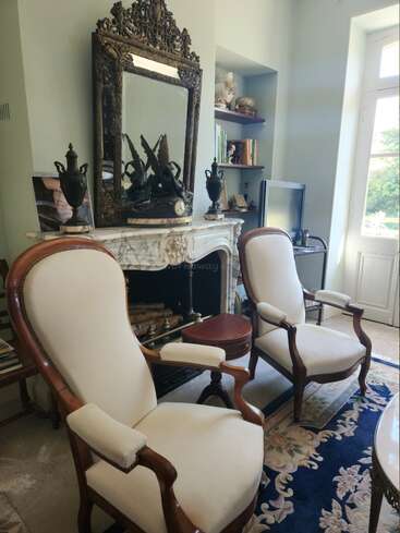 Dieses elegante Wohnzimmer verfügt über zwei antike Sessel, einen verzierten Spiegel über einem Marmorkamin, eine klassische Einrichtung, Bücherregale und ein großes Fenster, durch das Tageslicht hereinströmt.