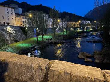 Ein ruhiger Fluss fließt in der Abenddämmerung durch eine bezaubernde Stadt, beleuchtet von Straßenlaternen und bunten Bodenleuchten, während sich die Berge als Silhouetten gegen den Abendhimmel abzeichnen. Eine friedliche Szenerie.