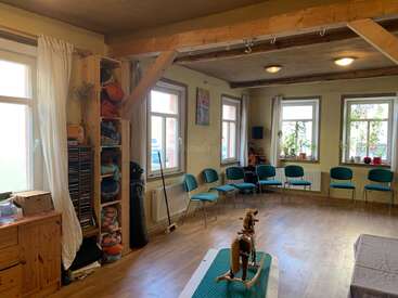 Una habitación acogedora y luminosa, con suelos y vigas de madera, sillas azules alrededor del perímetro, estanterías con mantas de colores, plantas y un caballito balancín como elemento central. Ambiente acogedor.