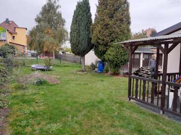 Un patio trasero cubierto de césped contiene un pequeño trampolín, árboles altos y arbustos. Una persona está de pie en un patio junto a unos tablones de madera. Se ven casas cercanas fuera de la valla.