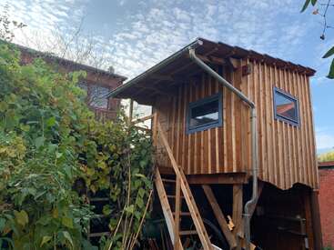 Una pequeña casa del árbol de madera se eleva con una escalera para acceder a ella. Rodeada de exuberante vegetación, cuenta con ventanas modernas, un techo inclinado y un canalón para la lluvia.