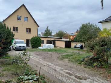 Una casa amarilla de tres pisos con una furgoneta aparcada al lado, un garaje, materiales de construcción esparcidos, algunos arbustos y un patio parcialmente cubierto de césped y parcialmente lleno de tierra.