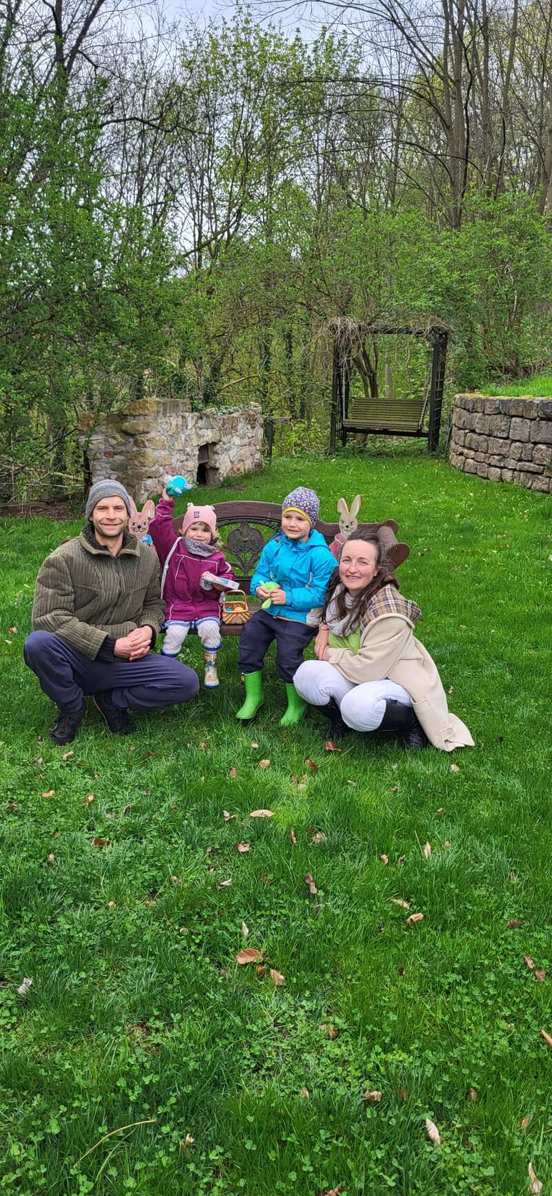 Una familia de cuatro miembros posa al aire libre en un jardín verde. Dos niños están sentados en un banco con cestas, flanqueados por sus padres sonrientes, rodeados de follaje primaveral.