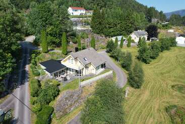 Uma pitoresca casa amarela é cercada por uma vegetação exuberante, árvores, estradas sinuosas e casas vizinhas, aninhada em uma paisagem rural tranquila sob um céu claro.