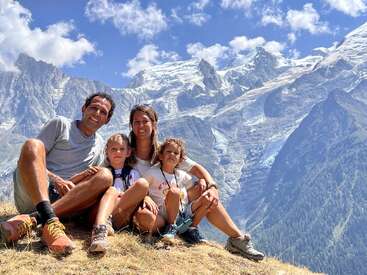 A imagem mostra uma família de quatro pessoas sentada em uma colina gramada, com uma majestosa cadeia de montanhas e um céu azul com nuvens como pano de fundo.