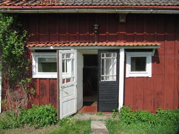A rustic red wooden house with a tiled roof, two white-trimmed windows, and an open split door; surrounded by greenery and exposed to bright sunlight.