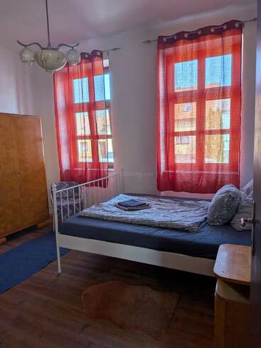 Un acogedor dormitorio con dos grandes ventanas, cortinas rojas, una cama de metal, ropa de cama estampada, un armario de madera, una lámpara de araña vintage, una mesita de noche y un cálido suelo de madera en tonos marrones.