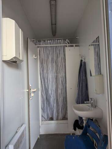 En esta imagen se ve un cuarto de baño pequeño con ducha, una cortina gris estampada, un lavabo, un espejo, toallas, una silla azul, un dispensador de toallas de papel y una lámpara fluorescente en el techo.