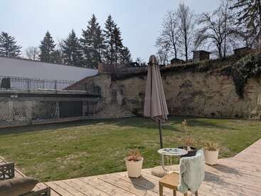 Una escena de un patio trasero con césped, una terraza de madera, plantas en macetas, una sombrilla cerrada, una mesa y una silla. Un muro de piedra y árboles al fondo. Ambiente tranquilo.