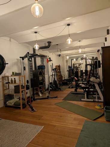 This image shows a cozy home gym with wooden floors, modern lighting, mirrors, exercise equipment, a yoga mat, shelves with towels, and various fitness accessories neatly arranged.