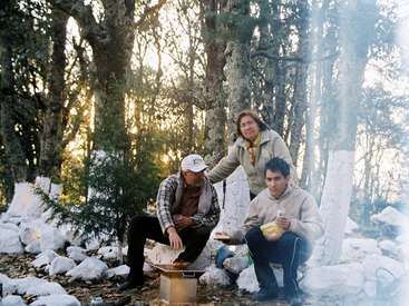 La imagen muestra a tres individuos en una zona boscosa, con dos hombres sentados sobre rocas y una mujer de pie detrás de ellos, rodeados de árboles y el suelo cubierto de nieve.