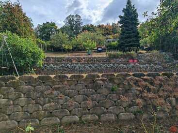A imagem mostra uma cena serena de jardim, com um muro de pedra, árvores exuberantes e plantas vibrantes, em um céu nublado com um toque de luz solar.
