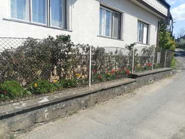 A imagem mostra uma casa branca com uma cerca de arame e um muro de contenção de concreto, com um jardim com flores e arbustos ao longo da calçada.
