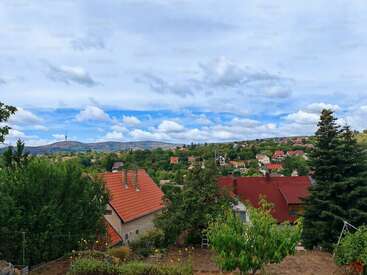 L'image représente un paysage serein composé de maisons, d'arbres et de montagnes sous un ciel nuageux, avec au loin une petite ville nichée dans les collines.