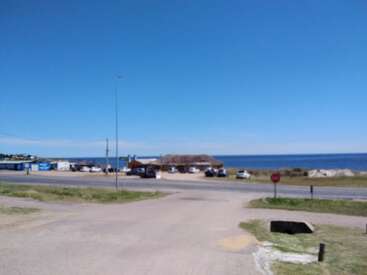 A bright, sunny coastal scene with a clear blue sky, ocean view, road, parked cars, thatched roof buildings, grassy areas, and a stop sign visible.